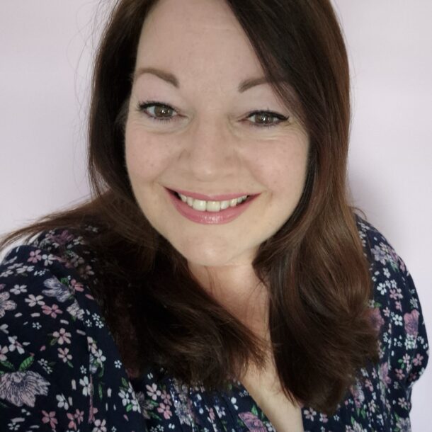 Jo Kelley A woman with long dark brown hair smiling in front of a light pink background, wearing a navy blue floral top with pink and purple flowers.