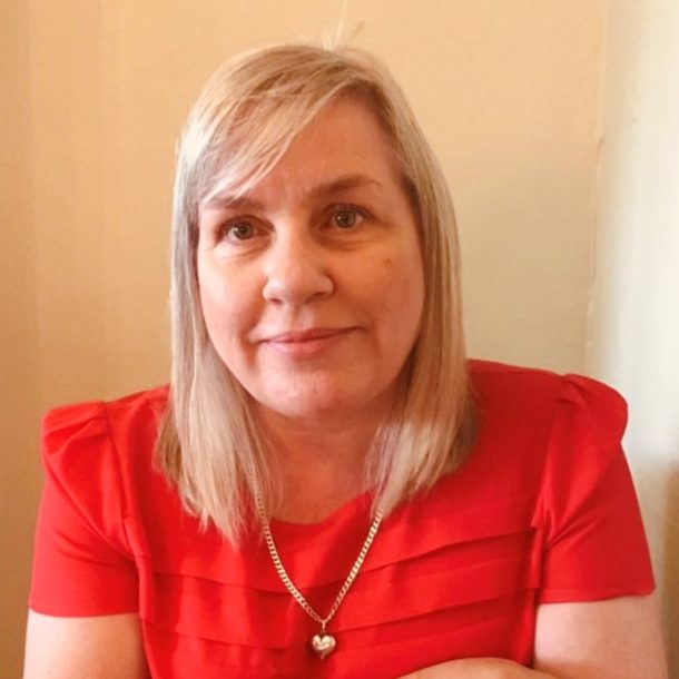 A person with light-colored, shoulder-length hair wearing a bright red top with pleated detailing and a gold heart-shaped pendant necklace, seated indoors against a neutral background.